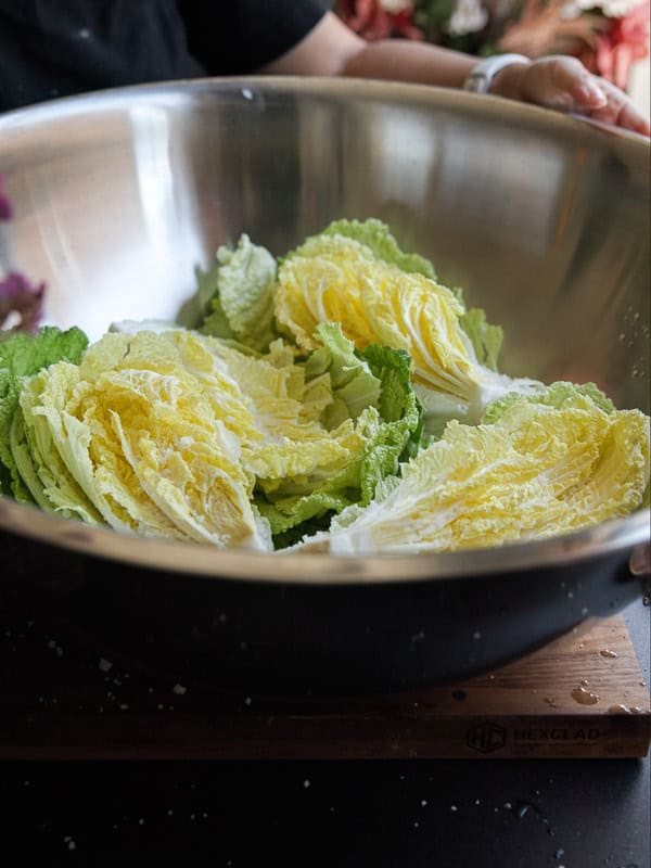 A large stainless steel bowl contains halved pieces of napa cabbage. The bowl is placed on a wooden cutting board, and a person is holding the edge of the bowl. The cabbage leaves are green and pale yellow.